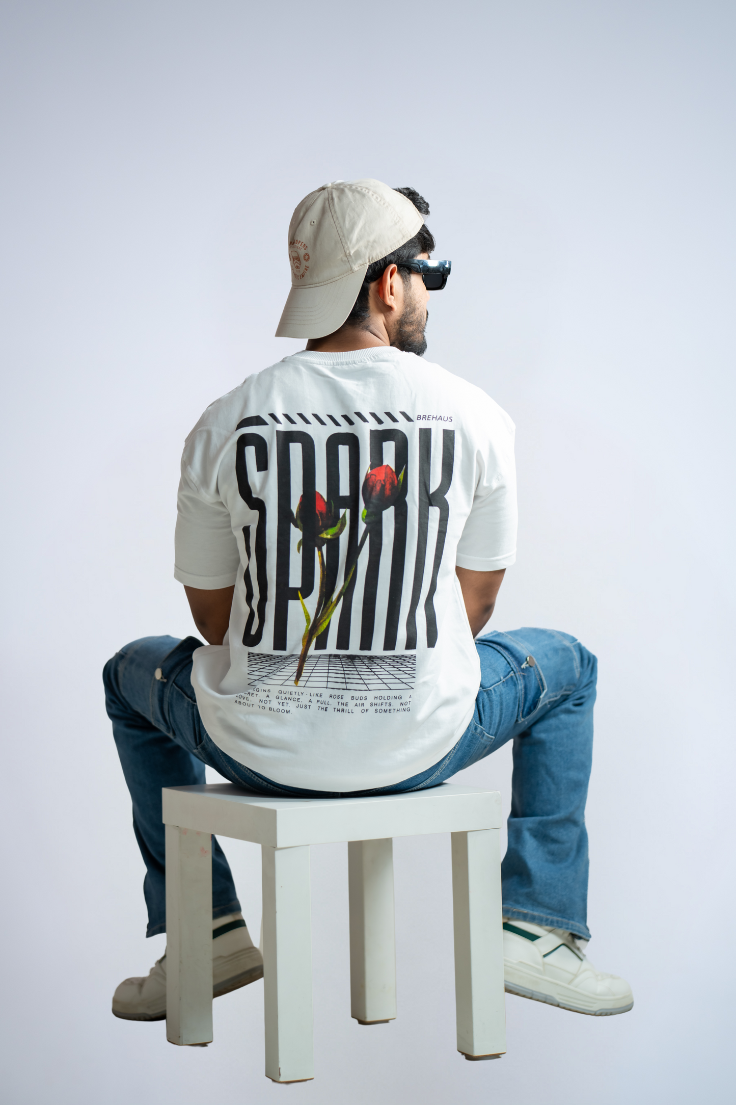 Person wearing a white t-shirt with 'SPARK' printed on the back, sitting on a white stool against a light gray background.