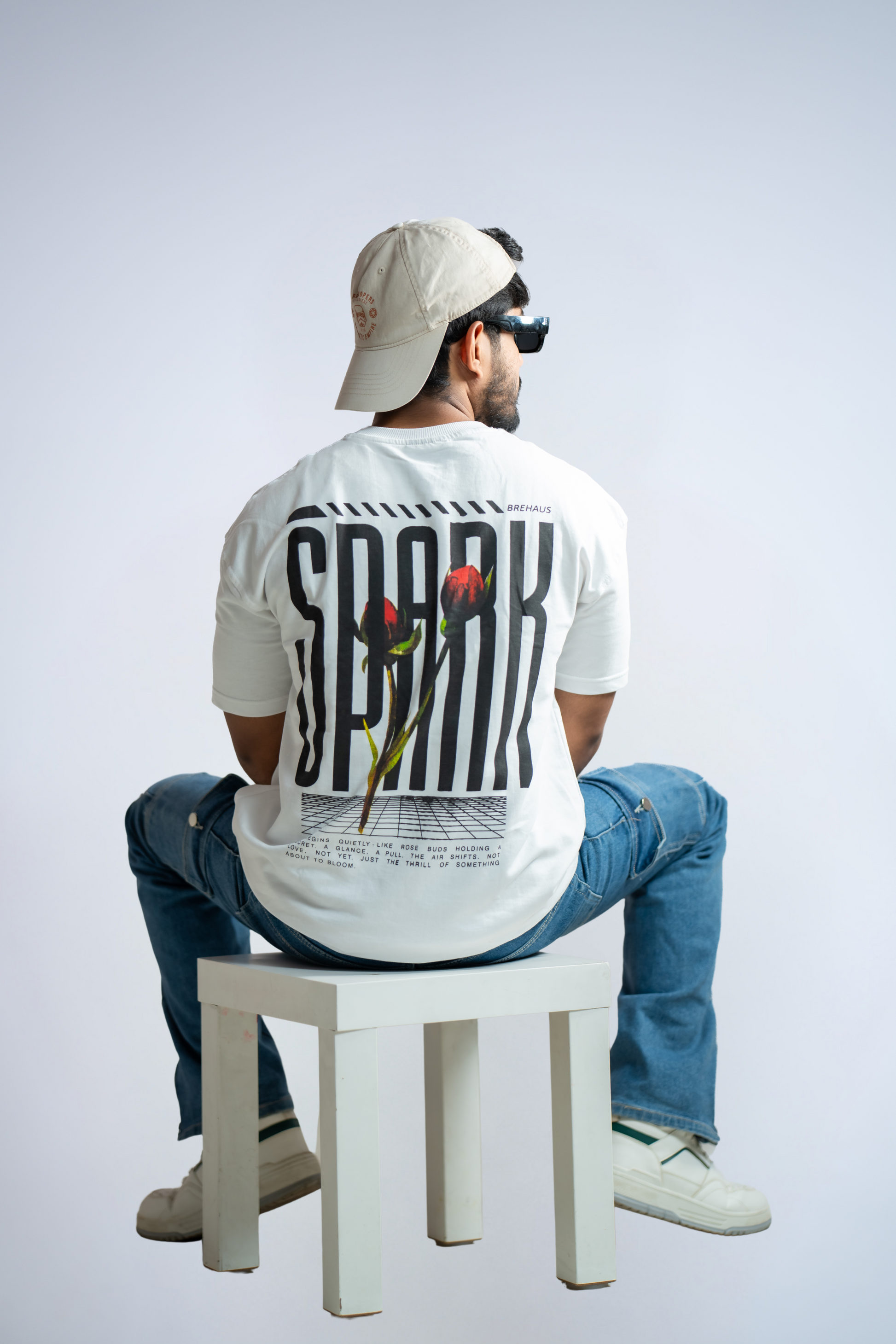 Person wearing a white t-shirt with 'SPARK' printed on the back, sitting on a white stool against a light gray background.